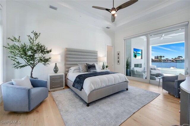 bedroom with access to outside, ceiling fan, light hardwood / wood-style floors, a raised ceiling, and a water view