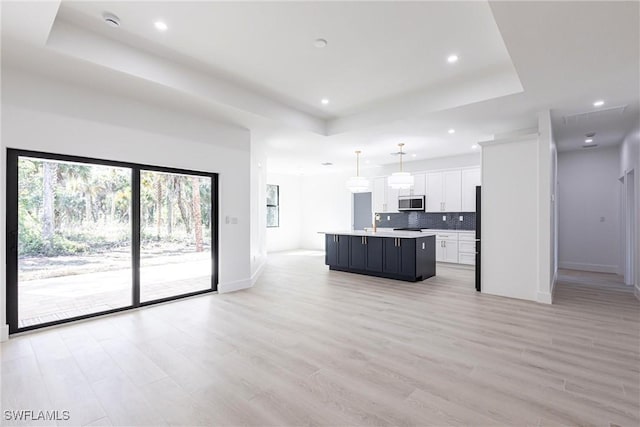 kitchen with pendant lighting, backsplash, a tray ceiling, an island with sink, and white cabinets