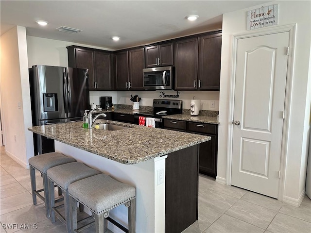 kitchen featuring appliances with stainless steel finishes, sink, light stone countertops, dark brown cabinets, and a center island with sink