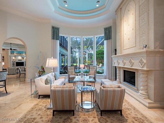 living area featuring crown molding, a premium fireplace, arched walkways, a raised ceiling, and a ceiling fan