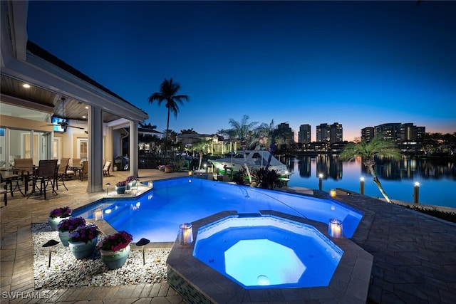 pool at dusk featuring an in ground hot tub, a water view, ceiling fan, and a patio