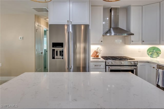 kitchen with white cabinetry, backsplash, light stone counters, premium appliances, and wall chimney exhaust hood