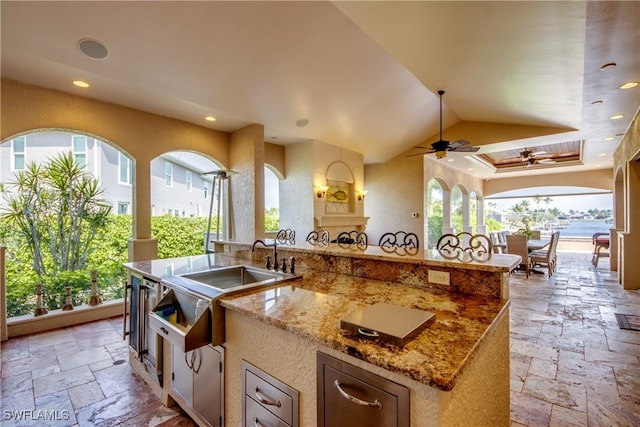 kitchen featuring ceiling fan, vaulted ceiling, a center island with sink, and light stone counters