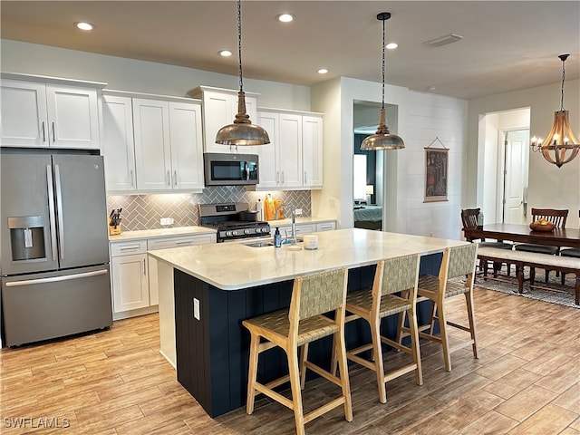 kitchen with stainless steel appliances, white cabinets, a center island with sink, light wood finished floors, and decorative light fixtures