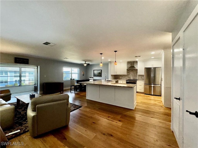 kitchen with wall chimney exhaust hood, white cabinetry, hanging light fixtures, stainless steel appliances, and a kitchen island with sink