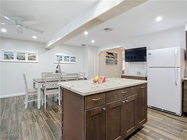 kitchen with a kitchen island, white refrigerator, ceiling fan, light stone counters, and dark brown cabinetry