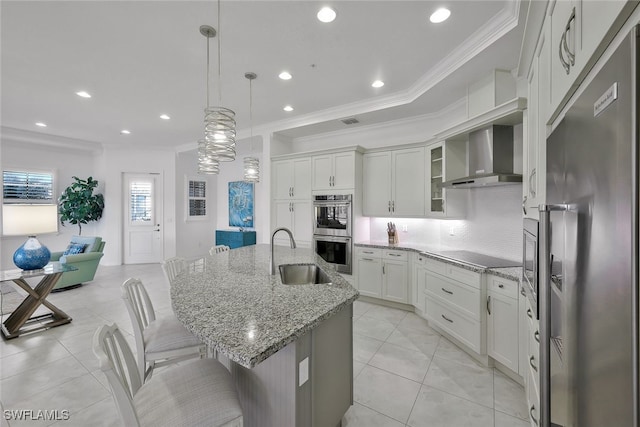 kitchen with sink, wall chimney range hood, appliances with stainless steel finishes, white cabinetry, and an island with sink