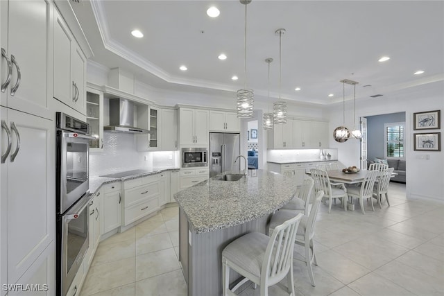 kitchen featuring wall chimney exhaust hood, light stone counters, hanging light fixtures, a center island with sink, and appliances with stainless steel finishes