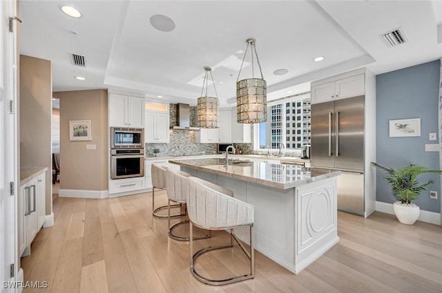 kitchen with built in appliances, a raised ceiling, white cabinets, and a center island with sink