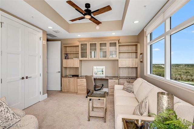 carpeted office featuring a raised ceiling, built in desk, and ceiling fan