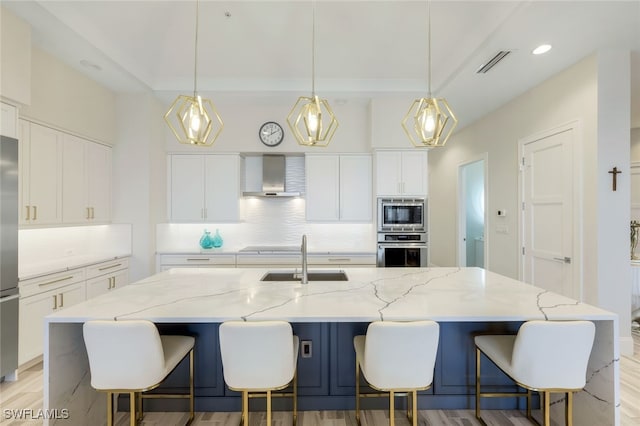 kitchen with a large island with sink, appliances with stainless steel finishes, wall chimney range hood, and hanging light fixtures