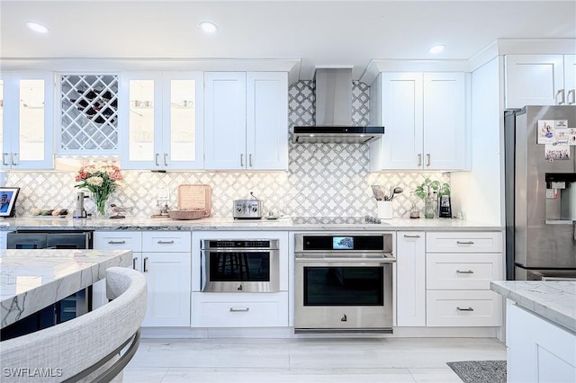 kitchen featuring wall chimney range hood, white cabinetry, stainless steel appliances, tasteful backsplash, and light stone countertops