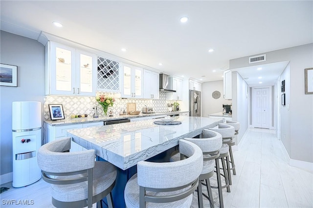 kitchen with wall chimney range hood, a breakfast bar area, stainless steel refrigerator with ice dispenser, light stone countertops, and white cabinets