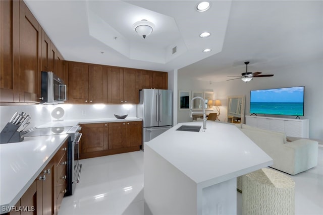 kitchen with a raised ceiling, an island with sink, sink, ceiling fan, and stainless steel appliances