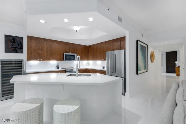 kitchen with a raised ceiling, an island with sink, sink, beverage cooler, and stainless steel appliances