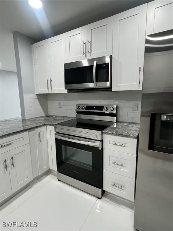 kitchen with light tile patterned floors, appliances with stainless steel finishes, white cabinetry, backsplash, and dark stone countertops