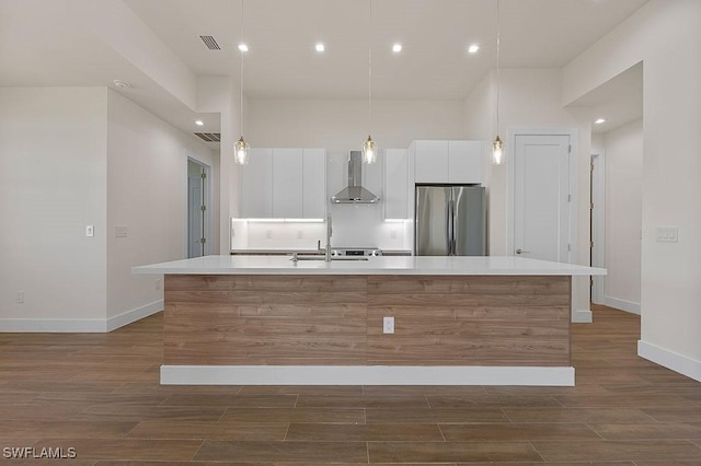 kitchen featuring stainless steel refrigerator, white cabinets, a kitchen island with sink, dark wood-type flooring, and wall chimney exhaust hood