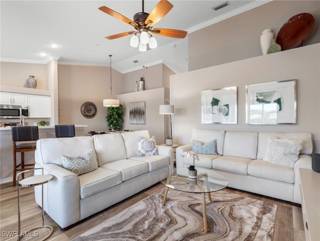 living room with visible vents, a towering ceiling, ceiling fan, crown molding, and light wood-type flooring