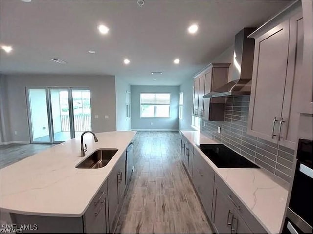 kitchen with sink, wall chimney range hood, light stone countertops, black electric cooktop, and a large island with sink