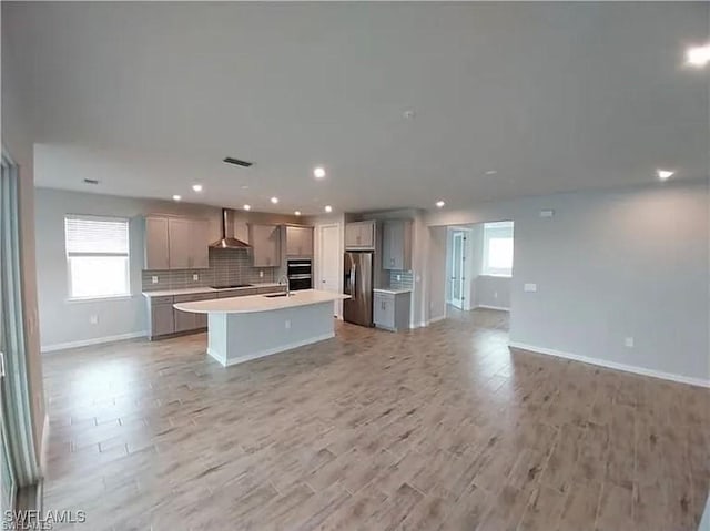 kitchen featuring appliances with stainless steel finishes, gray cabinetry, backsplash, wall chimney range hood, and a center island with sink