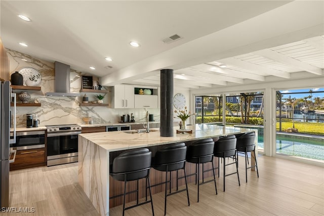 kitchen featuring light stone counters, appliances with stainless steel finishes, an island with sink, white cabinets, and wall chimney range hood