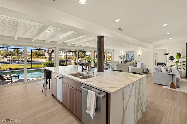 kitchen featuring sink, an island with sink, stainless steel dishwasher, beamed ceiling, and light wood-type flooring