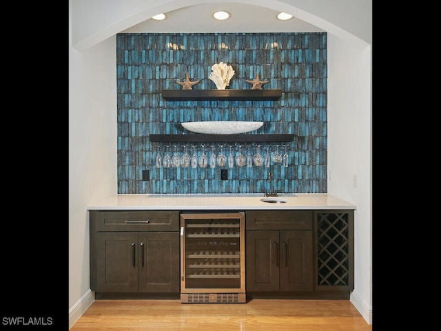 bar with wine cooler, decorative backsplash, dark brown cabinets, and light wood-type flooring