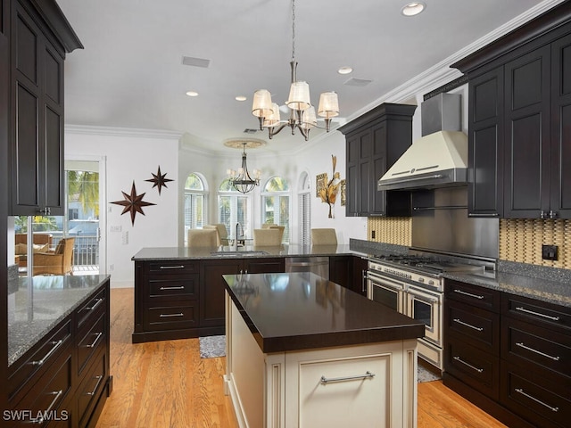 kitchen featuring pendant lighting, an inviting chandelier, stainless steel appliances, light wood-type flooring, and wall chimney exhaust hood