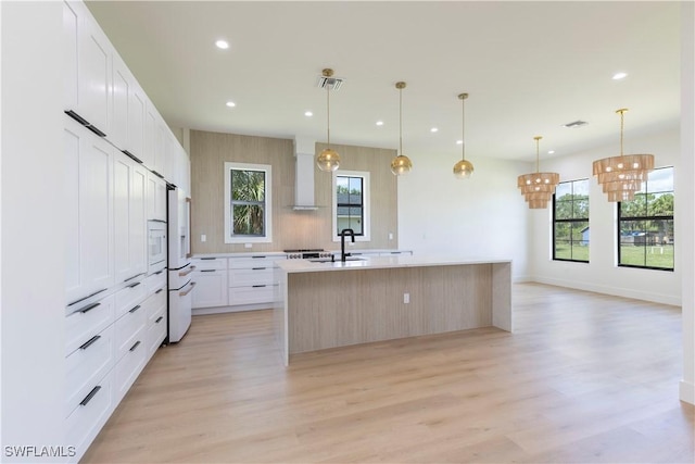 kitchen featuring wall chimney range hood, light hardwood / wood-style flooring, a kitchen island with sink, white cabinetry, and decorative light fixtures