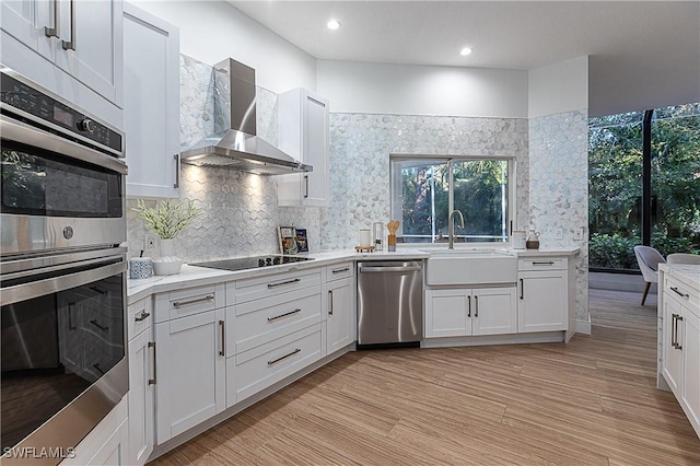 kitchen with wall chimney exhaust hood, sink, light wood-type flooring, appliances with stainless steel finishes, and white cabinets