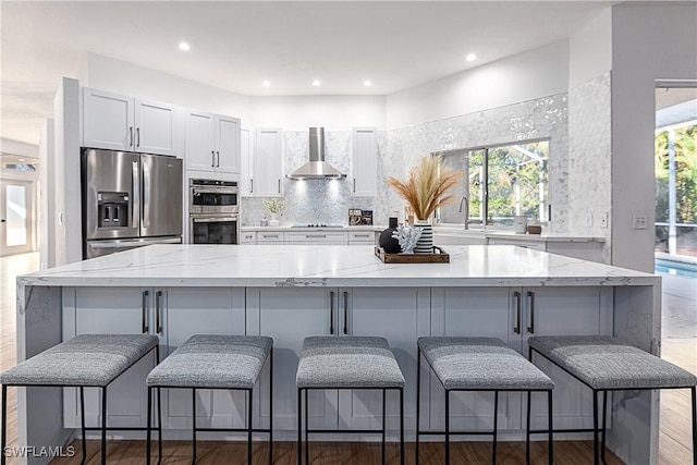 kitchen with a breakfast bar, white cabinetry, stainless steel appliances, light stone counters, and wall chimney exhaust hood