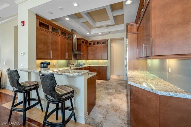 kitchen with a breakfast bar area, coffered ceiling, light stone counters, kitchen peninsula, and wall chimney exhaust hood