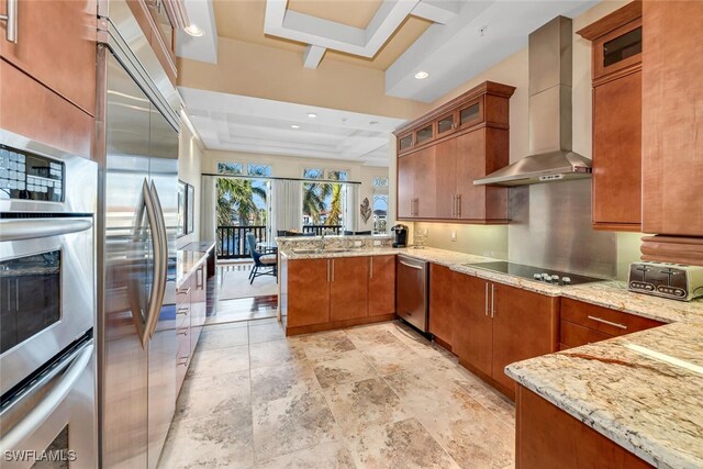 kitchen with appliances with stainless steel finishes, kitchen peninsula, a raised ceiling, light stone countertops, and wall chimney exhaust hood