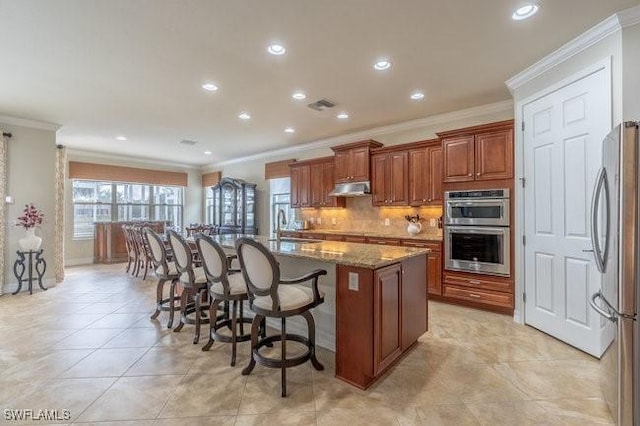 kitchen with a kitchen island with sink, backsplash, stainless steel appliances, a kitchen bar, and dark stone counters