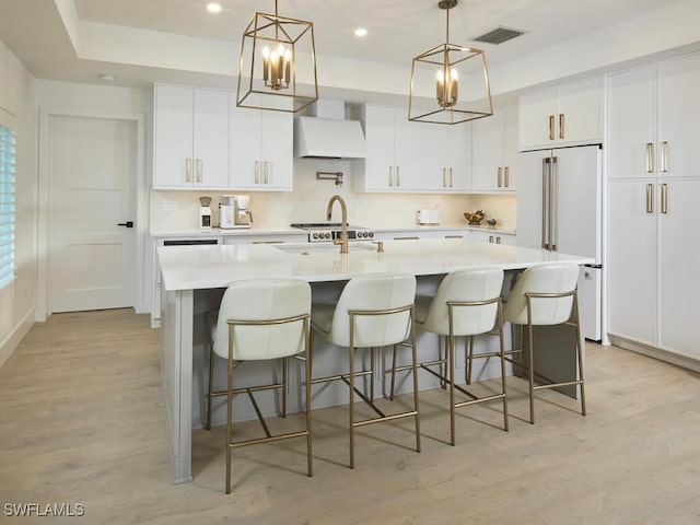 kitchen featuring an island with sink, high end white fridge, hanging light fixtures, and white cabinets