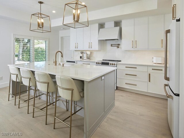 kitchen featuring a large island, wall chimney range hood, white appliances, white cabinetry, and decorative light fixtures