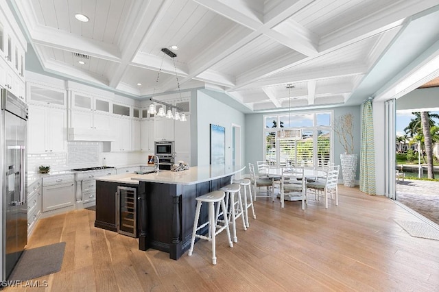 kitchen with built in appliances, hanging light fixtures, a center island with sink, beverage cooler, and white cabinets