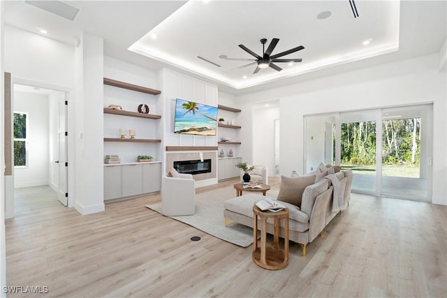 living area with a raised ceiling, ceiling fan, light wood finished floors, and a glass covered fireplace