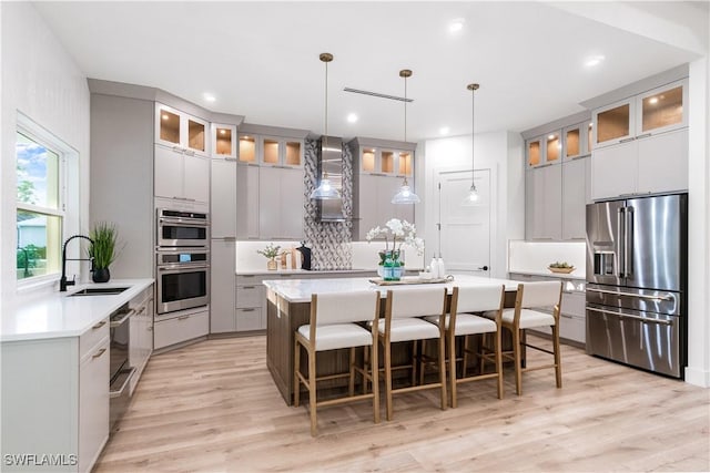 kitchen featuring a sink, stainless steel appliances, wall chimney exhaust hood, an island with sink, and gray cabinets