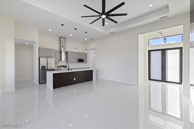 kitchen with wall chimney range hood, gray cabinets, a kitchen island with sink, stainless steel appliances, and decorative light fixtures