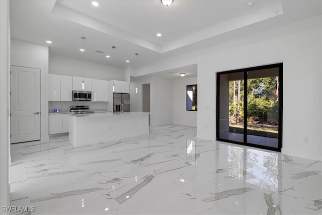 kitchen with a raised ceiling, white cabinets, hanging light fixtures, a kitchen island with sink, and stainless steel appliances