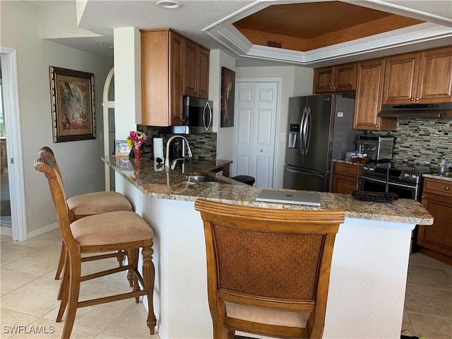 kitchen with light tile patterned floors, under cabinet range hood, stainless steel appliances, a kitchen breakfast bar, and a tray ceiling