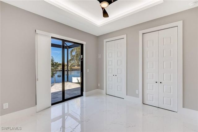 unfurnished bedroom with access to exterior, ceiling fan, a tray ceiling, multiple closets, and a water view