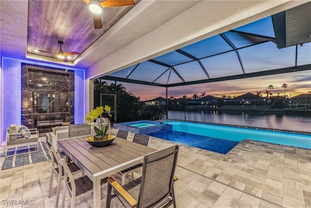 pool at dusk featuring ceiling fan, a water view, glass enclosure, a patio area, and an in ground hot tub