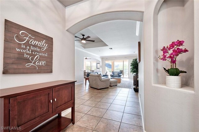 corridor with arched walkways, a raised ceiling, ornamental molding, light tile patterned flooring, and baseboards