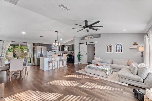 living room with dark wood-type flooring, a barn door, ceiling fan with notable chandelier, and vaulted ceiling