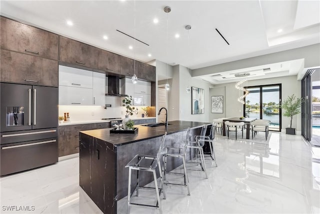 kitchen with sink, a center island with sink, paneled refrigerator, white cabinets, and wall chimney range hood