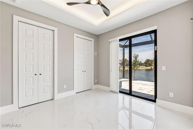 unfurnished bedroom featuring ceiling fan, access to exterior, a water view, two closets, and a raised ceiling