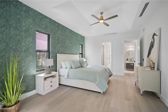 bedroom featuring ceiling fan, ensuite bath, a tray ceiling, and light wood-type flooring