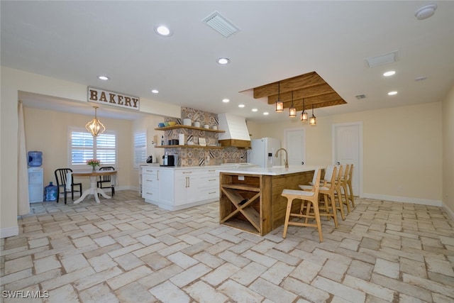 kitchen featuring premium range hood, white cabinetry, hanging light fixtures, white fridge with ice dispenser, and an island with sink